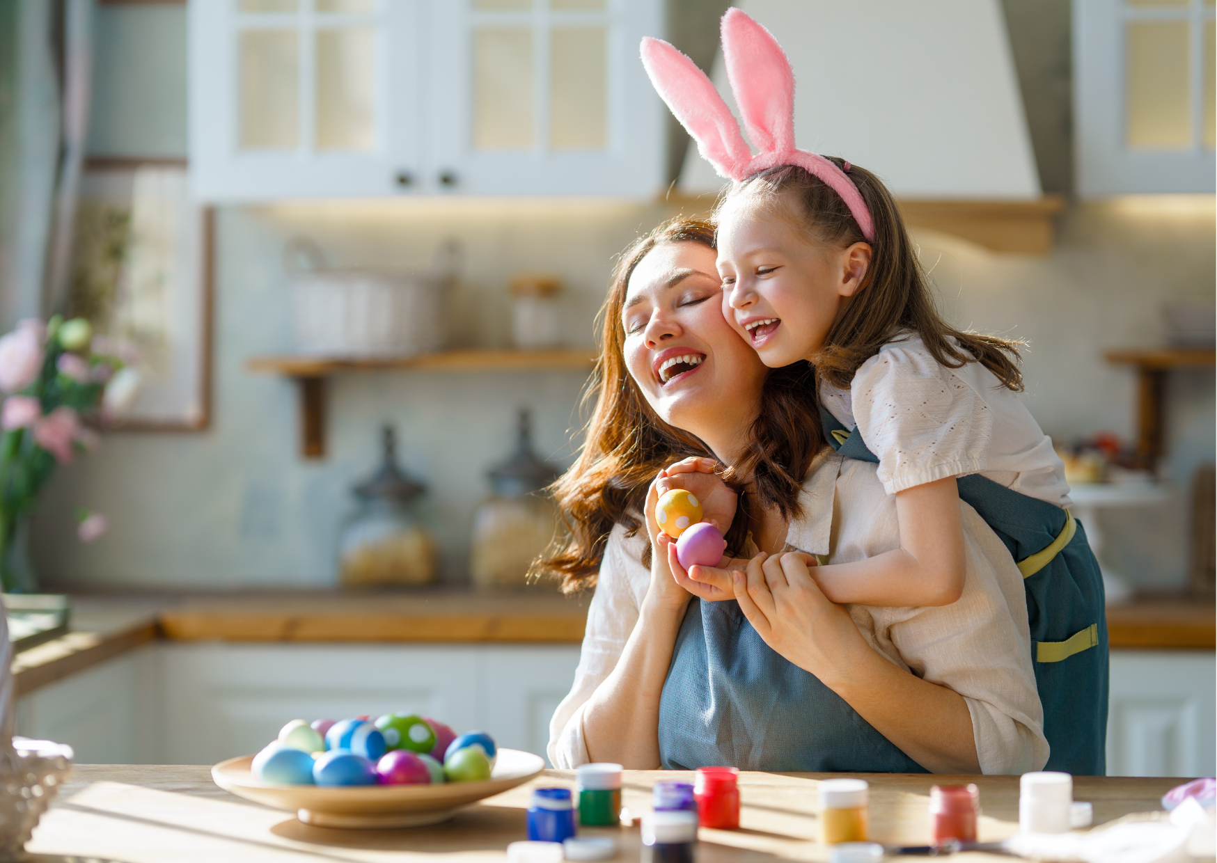Mother and daughter joyfully painting Easter eggs together in a sunlit kitchen.