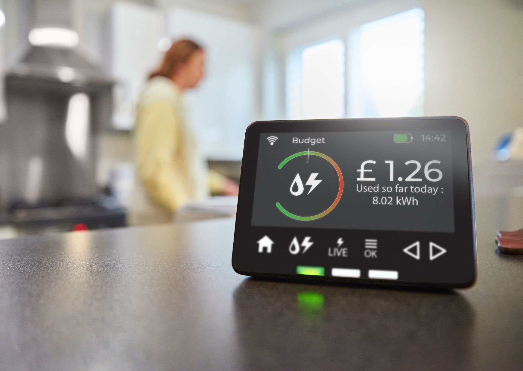 Image show smart meter in foreground. Lady wearing yellow top is blurred in the background. She is in what looks like a kitchen.
