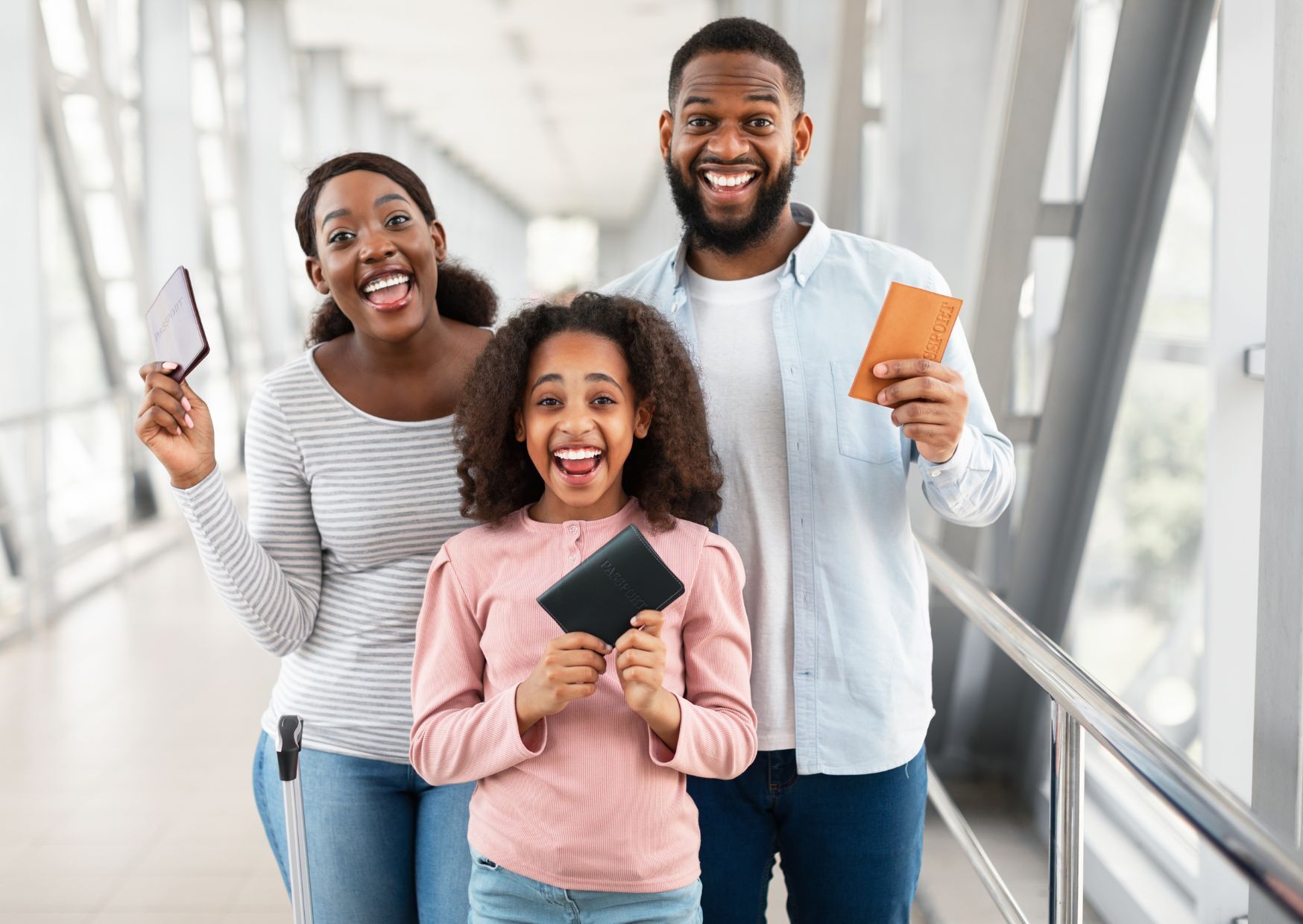 A woman, man, and child all with dark hair, smiling, wearing casual clothing, carrying passports. The background is an airport gangway.