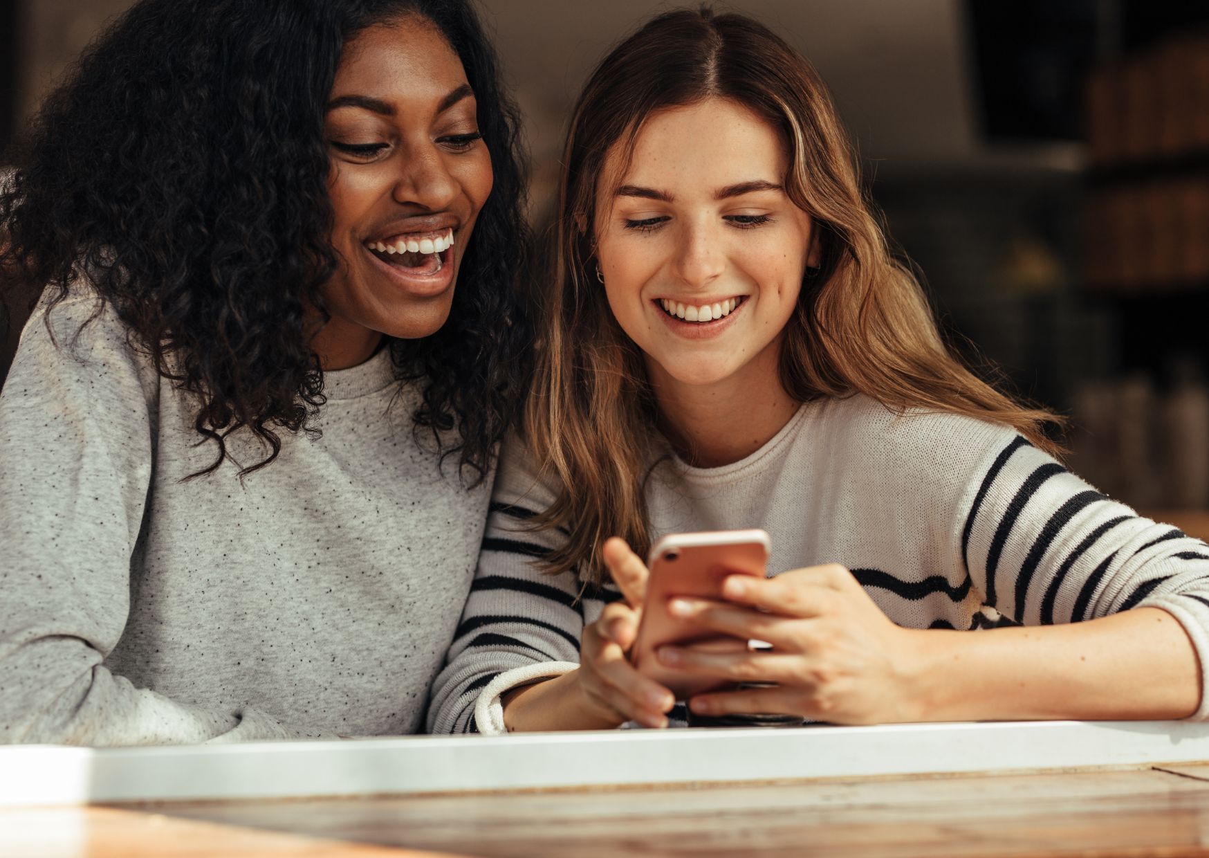 Two women, one with dark hair, one fair. Both wearing jumpers, looking a phone, delighted.