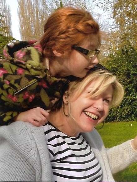 Image shows woman with light hair wearing striped tee shirt balancing young girl with red hair and glasses on her back. Trees in the backgroun.