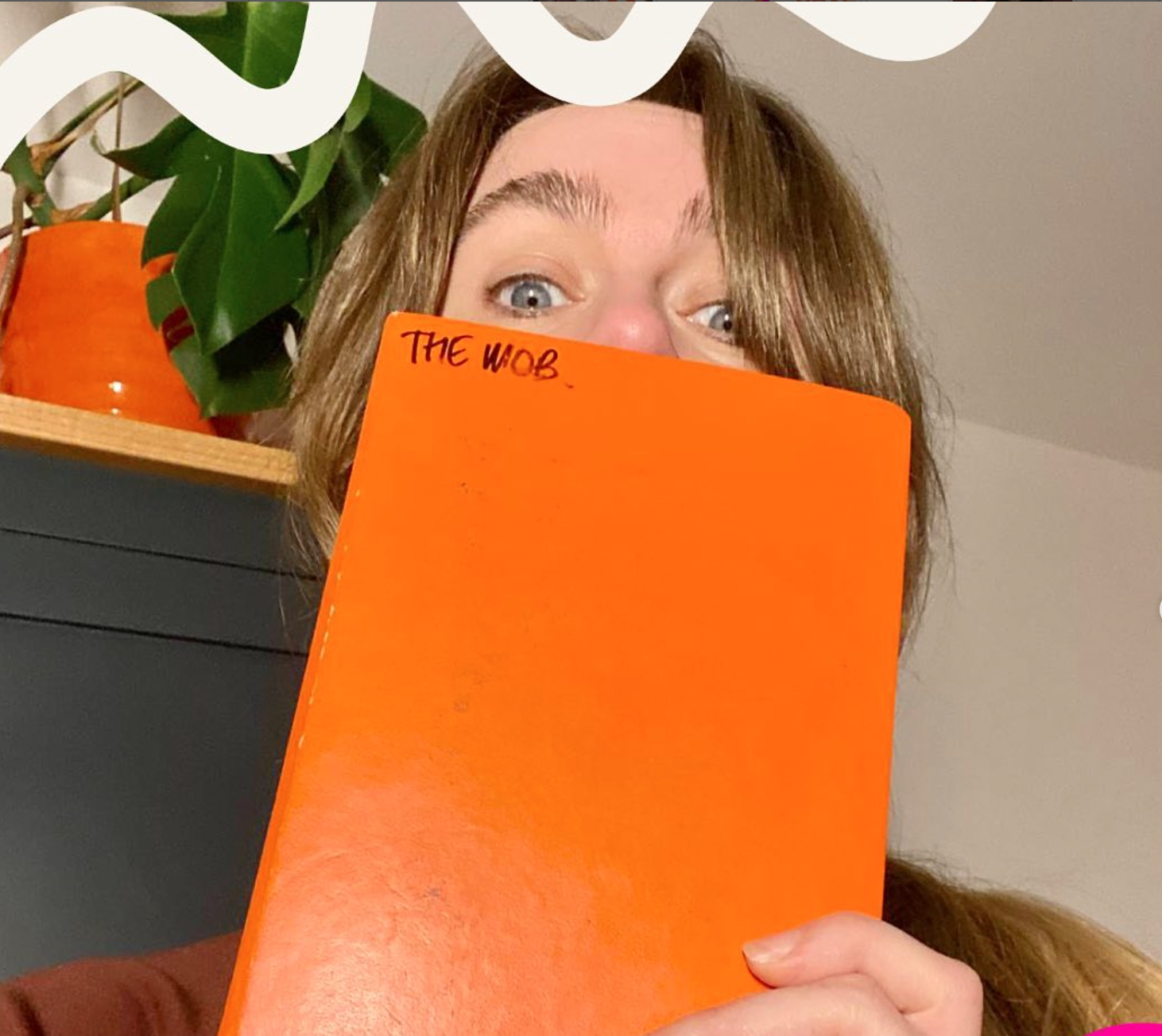 Image shows woman with brown hair peeking over a bright orange note book.