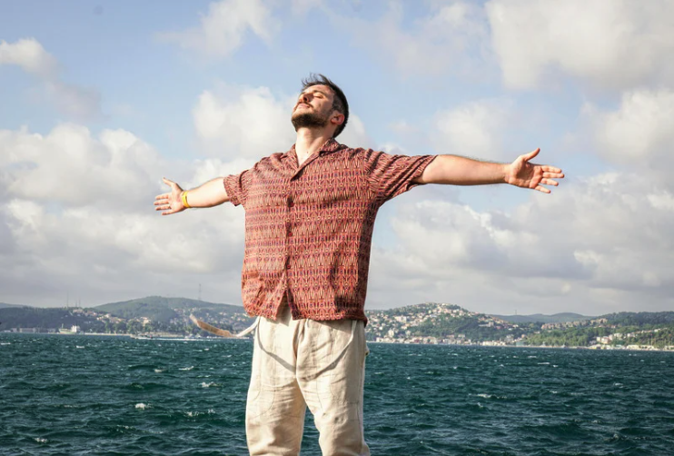 Image shows man with brown hair, and beard. He is standing with his arms open wide, looking up to the sky. He is wearing a burgandy short sleeved shirt, and light chinos.  In the background there is what looks like choppy waves and a hilly coastline.